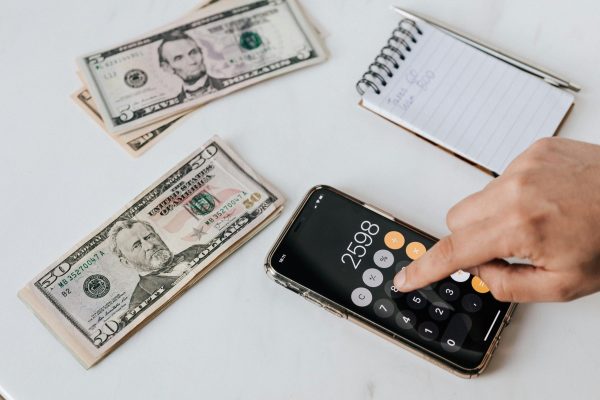 pexels-photo-4386324-4386324 From above of crop anonymous economist calculating total amount of income using calculator app on cellphone near piles of greenbacks and notebook with pen