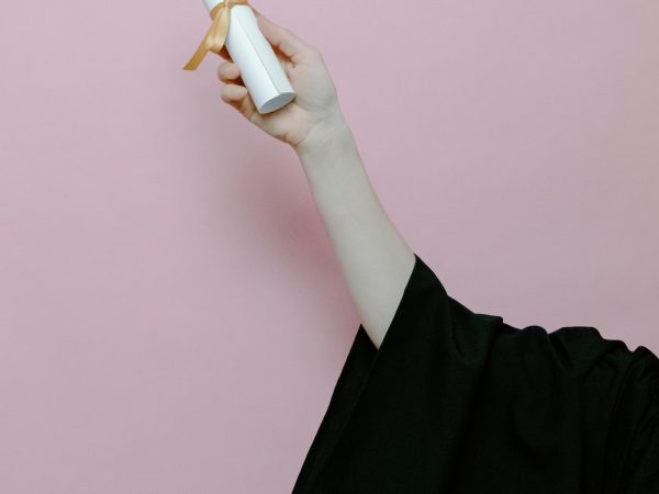 pexels-photo-7723833-7723833 Hand of a graduate wearing a gown holding a diploma with a pink background.